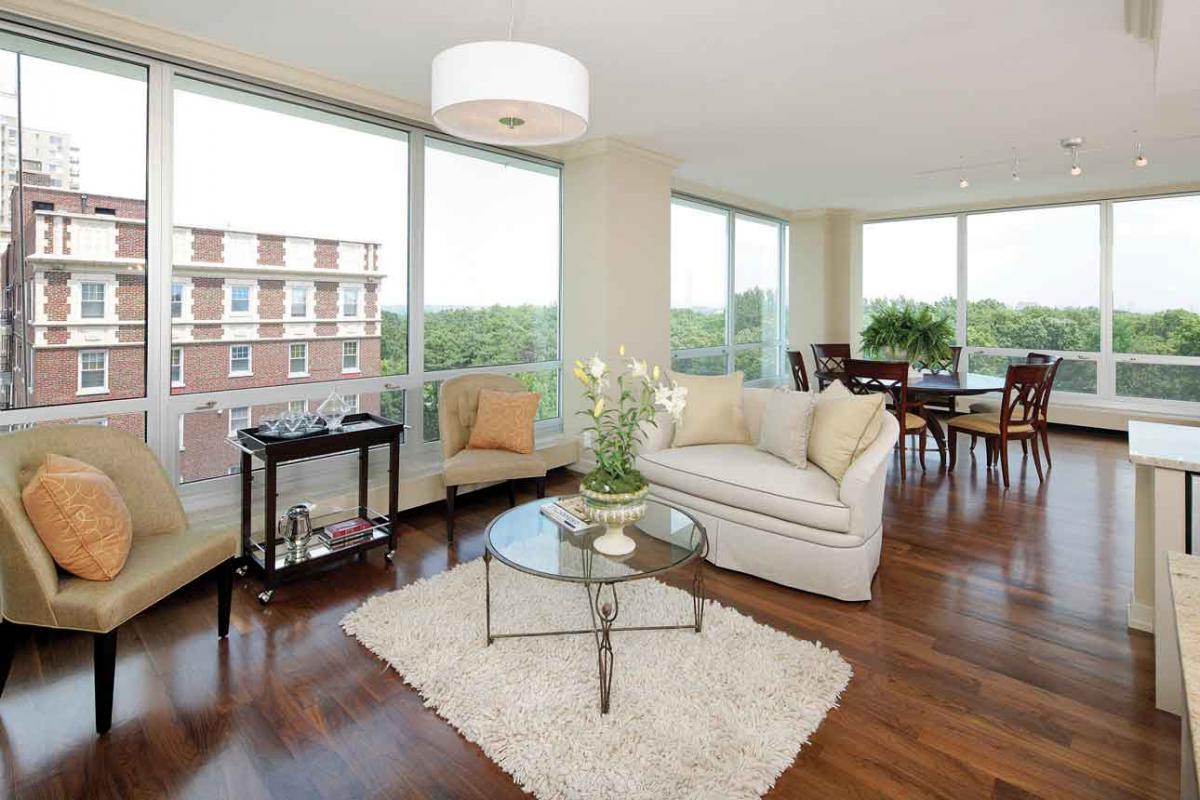This living room area has window walls, a white sofa, two tan chairs, a white rug, a black bar cart and a glass coffee table with flowers on it.