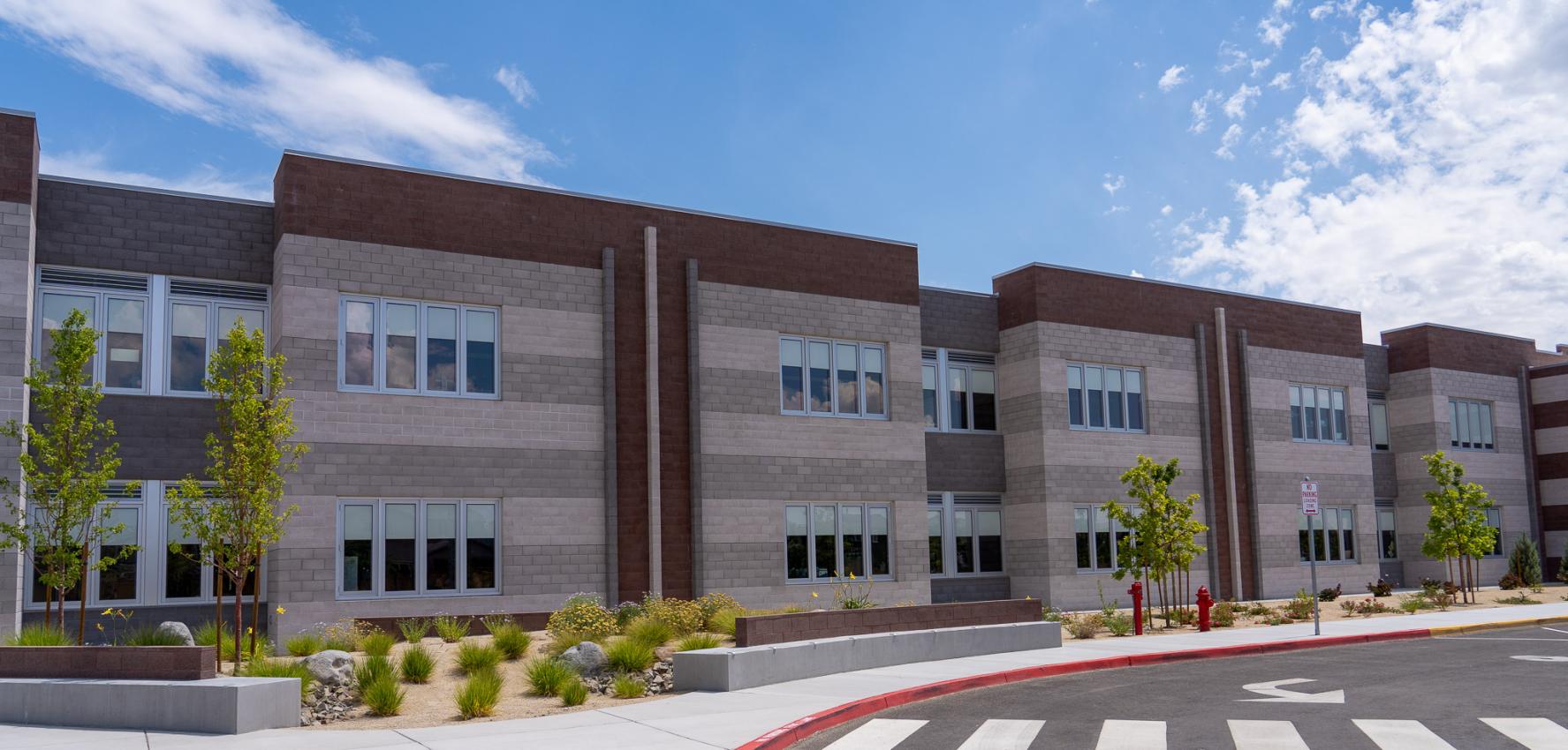 A two-story school building with brick in shades of gray and tan. The sidewalk in front is landscaped with small green bushes and trees.