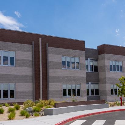 A two-story school building with brick in shades of gray and tan. The sidewalk in front is landscaped with small green bushes and trees.
